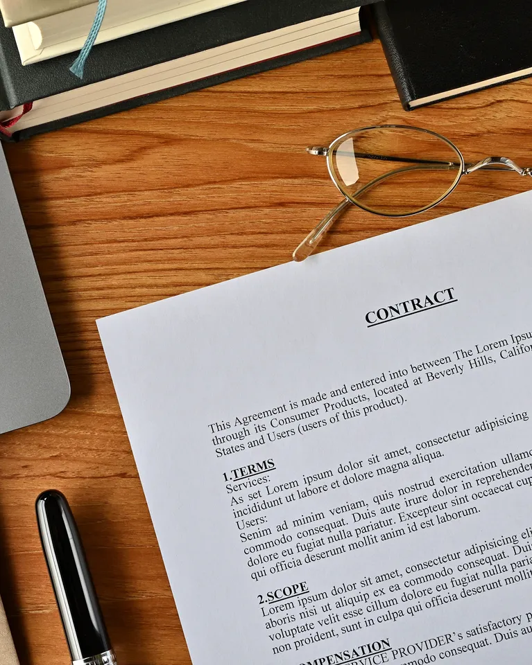 Top view of a wooden desk with a contract document, eyeglasses, pen, books, and a laptop corner.