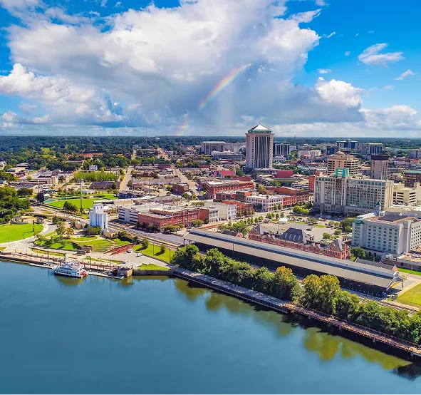 Aerial view of a cityscape with a river, green parks, buildings, and a faint rainbow under a partly cloudy sky.