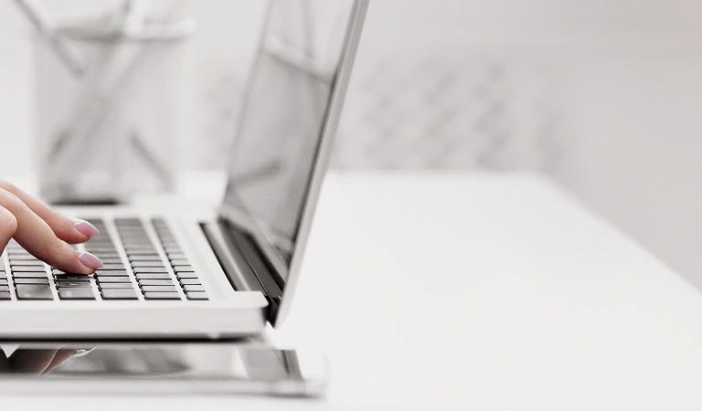 Close-up of a hand typing on a silver laptop keyboard on a white desk.